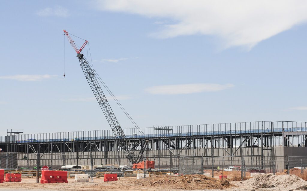 Construction site of a data center with a large crane lifting materials. The steel framework of the building is visible, with workers and vehicles operating in the background.