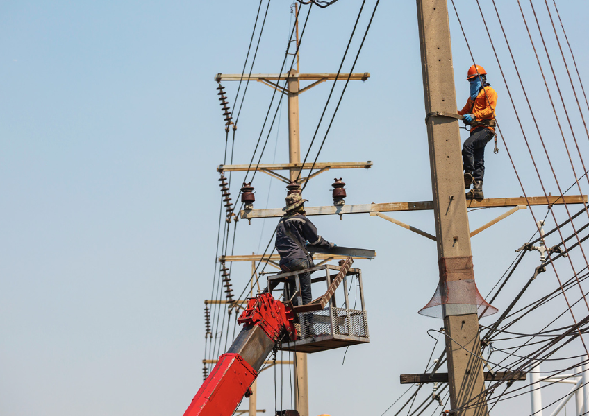 Two utility workers repairing or installing power lines—one elevated in a lift and another climbing a pole—against a clear blue sky
