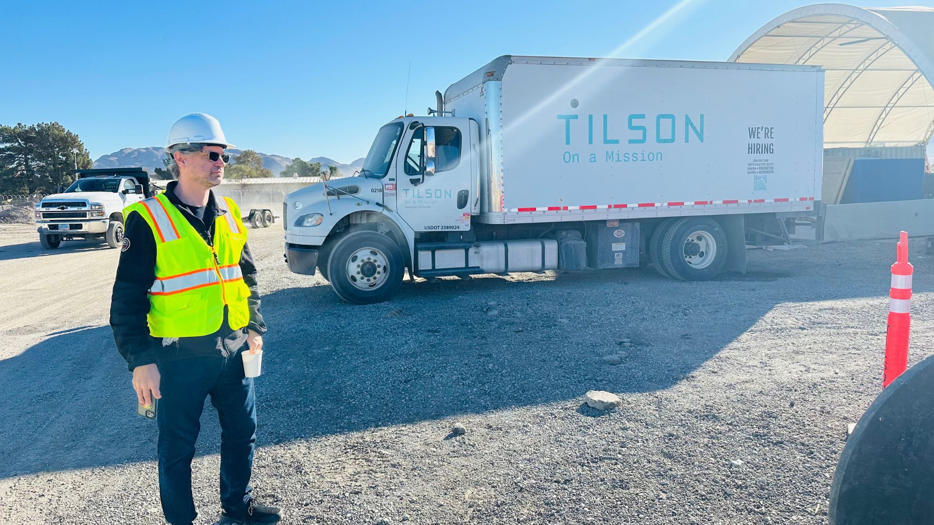 A construction worker wearing a white hard hat, sunglasses, and a high-visibility yellow safety vest stands in a gravel lot holding a coffee cup and a smartphone. Behind him, a large white Tilson-branded truck with the slogan "On a Mission" is parked, along with another white utility vehicle.
